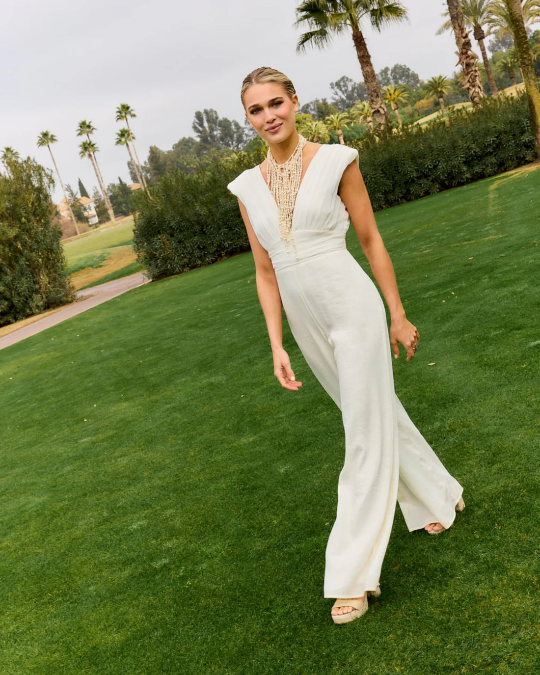 Woman wearing white Carlotta Jumpsuit on grass with palm trees — Lady Pipa | Endless UAE
