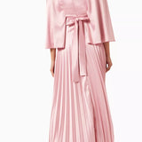 Woman wearing a pink satin pleated dress with matching cape overlay and metallic heels — Nass | Endless UAE