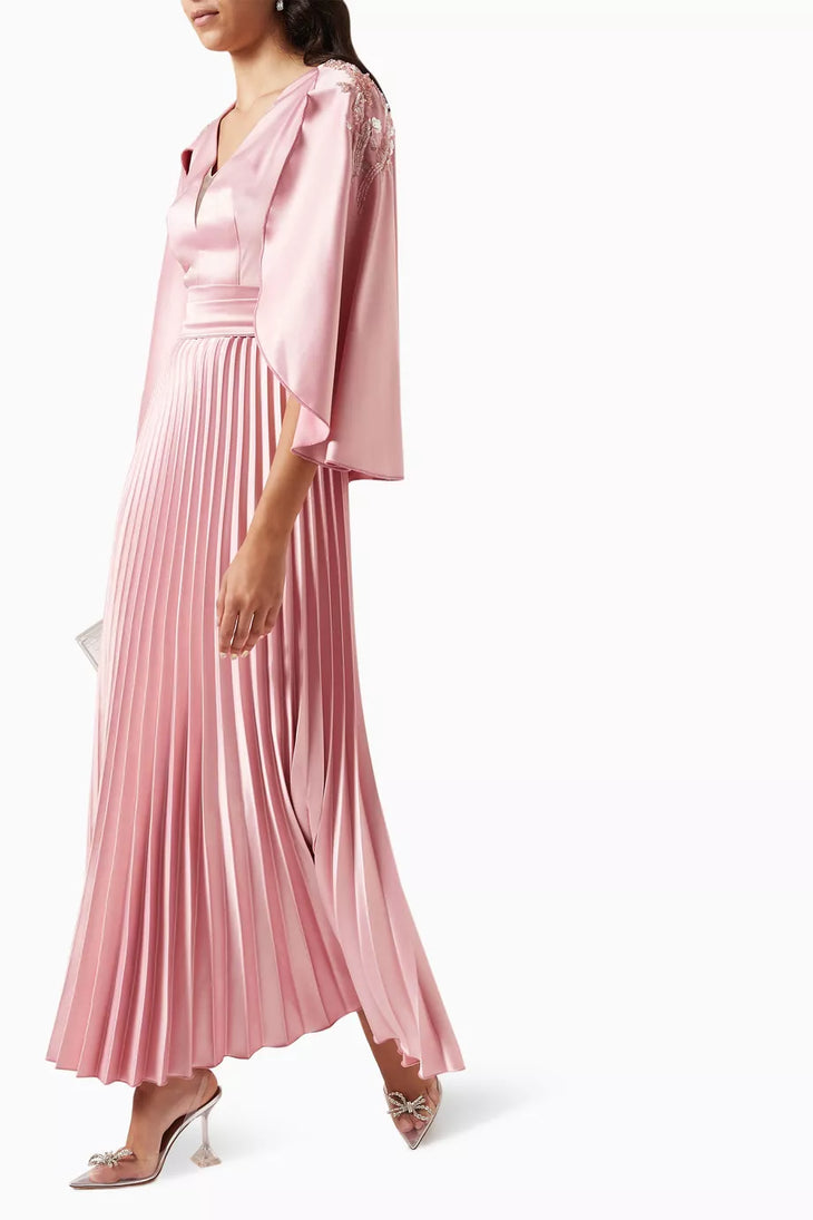 Woman wearing a pink satin pleated dress with matching cape overlay and metallic heels — Nass | Endless UAE