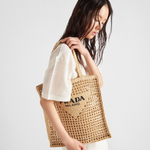 Natural raffia woven tote bag with black triangle logo, carried on shoulder by model in white outfit — Prada | Endless UAE