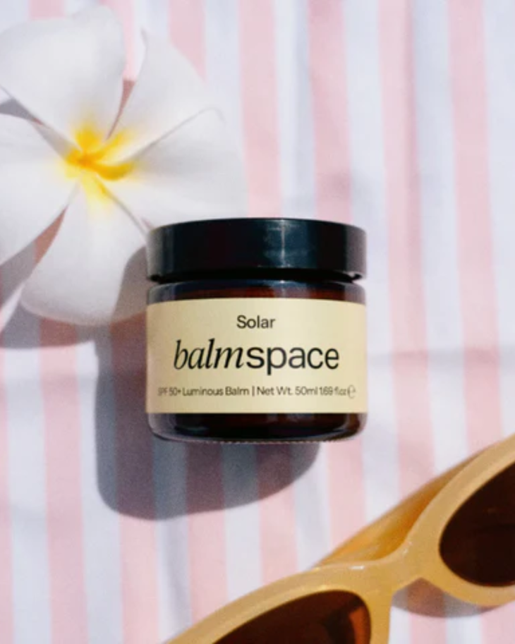 SPF 50+ luminous face balm in amber glass jar with cream label and black lid on pink striped background with plumeria flower — Balm Space | Endless UAE