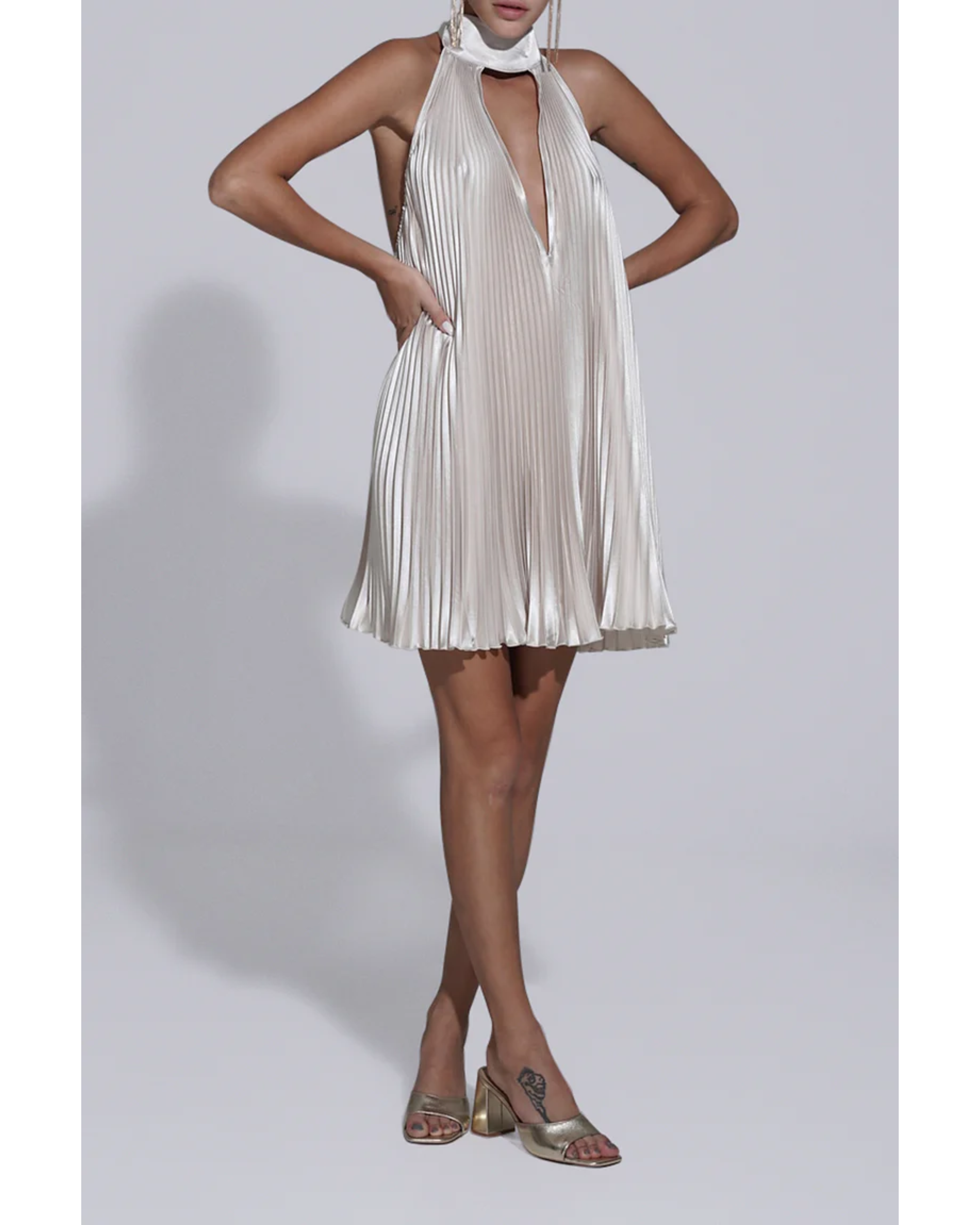 Woman wearing champagne pleated halterneck mini dress with deep V-neckline and metallic heeled sandals — L'IDÉE | Endless UAE