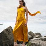 Woman wearing golden yellow silk slip dress standing on rocks by the sea — Vasiliki | Endless UAE
