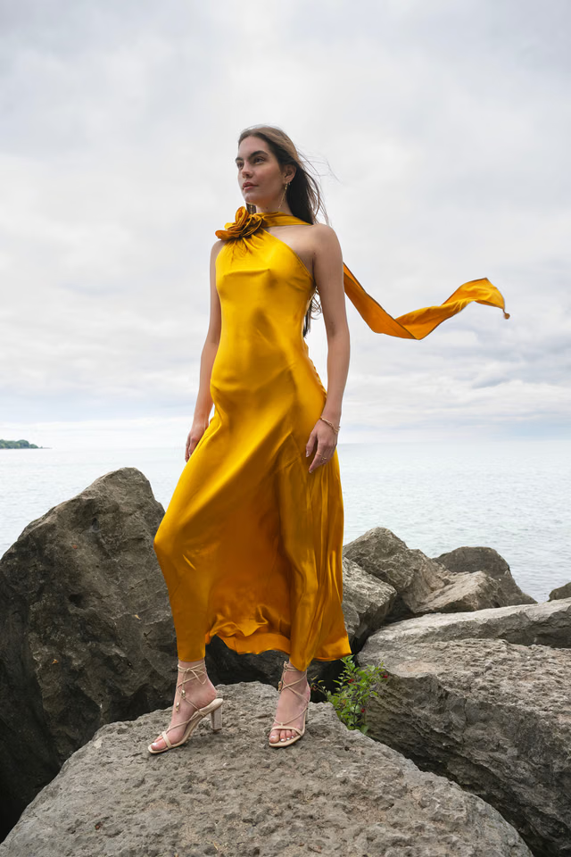 Woman wearing golden yellow silk slip dress standing on rocks by the sea — Vasiliki | Endless UAE