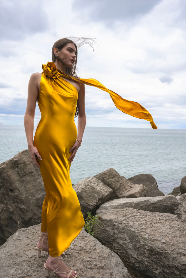 Woman wearing golden yellow silk slip dress standing on rocks by the sea — Vasiliki | Endless UAE