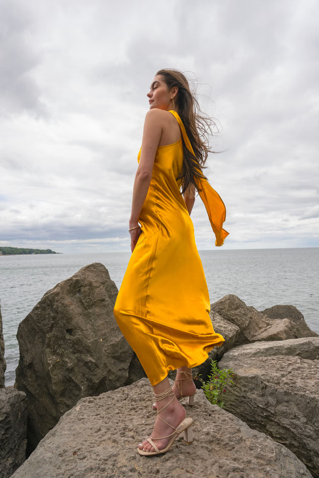 Woman wearing golden yellow silk slip dress standing on rocks by the sea — Vasiliki | Endless UAE