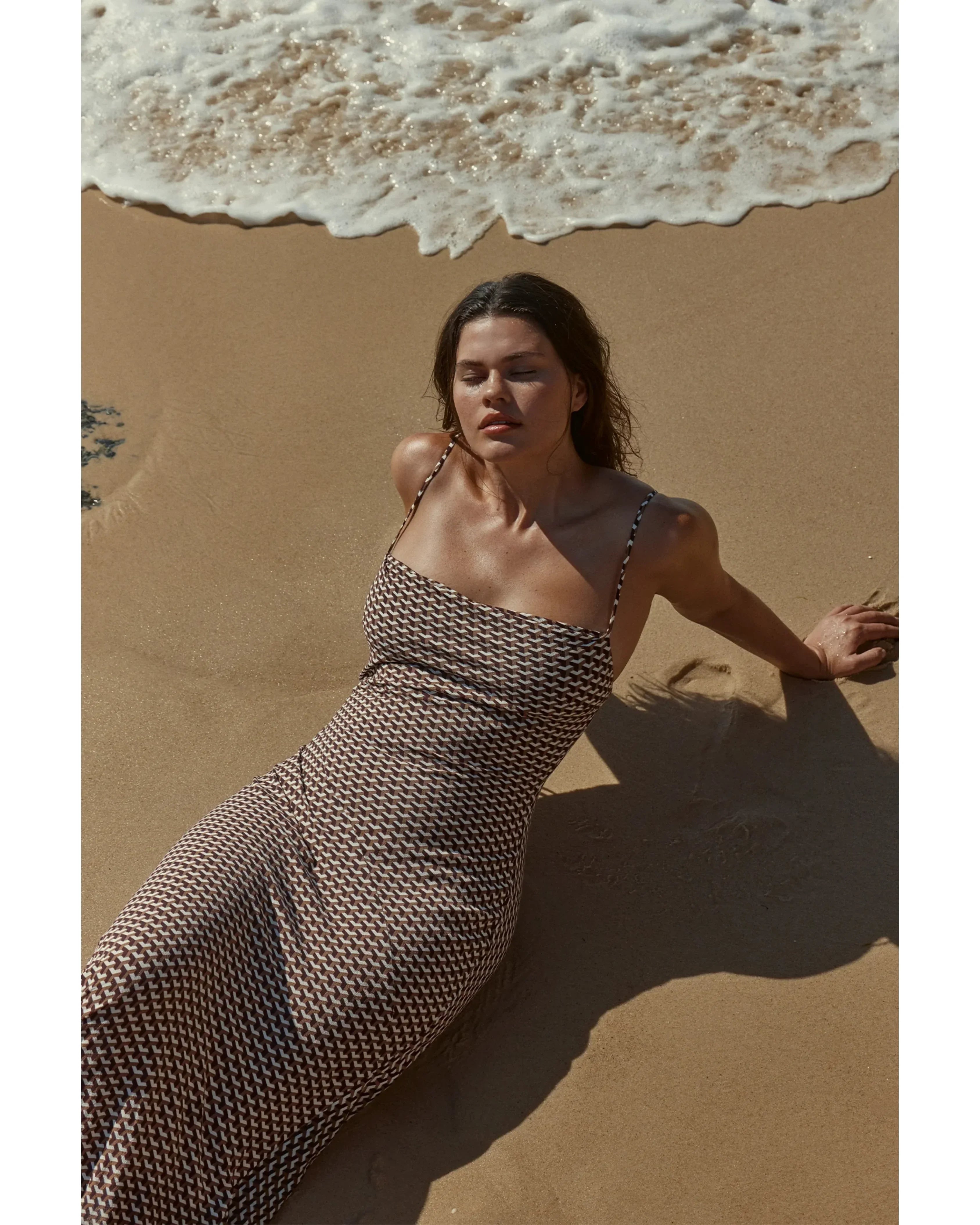 Woman in patterned maxi dress relaxing on beach sand with ocean waves — Monday Swimwear | Endless UAE