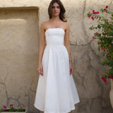 Woman in white strapless midi dress with pleated skirt and white heels against rustic wall — Cleopatress | Endless UAE