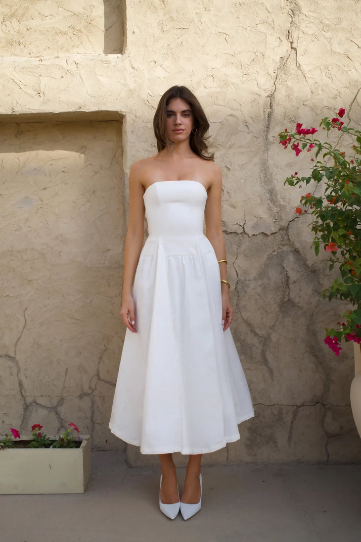 Woman in white strapless midi dress with pleated skirt and white heels against rustic wall — Cleopatress | Endless UAE