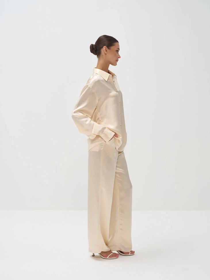 Woman in profile wearing cream silk pajama set with long sleeve button-up shirt and wide-leg pants — Nora The Label | Endless UAE