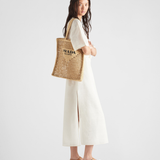 Woman carrying natural raffia woven tote bag with black Prada logo, wearing cream dress and sandals — Prada | Endless UAE