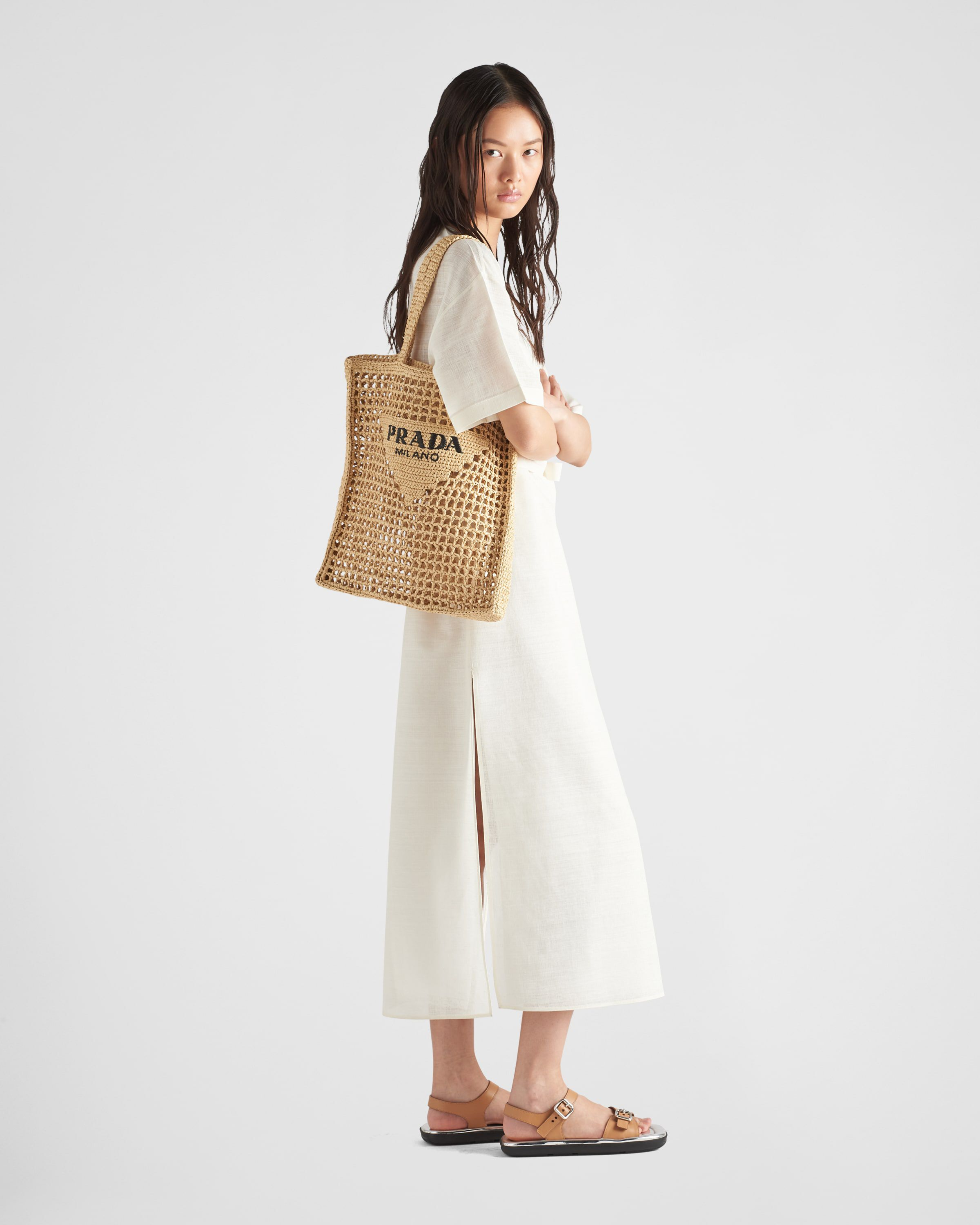 Woman carrying natural raffia woven tote bag with black Prada logo, wearing cream dress and sandals — Prada | Endless UAE