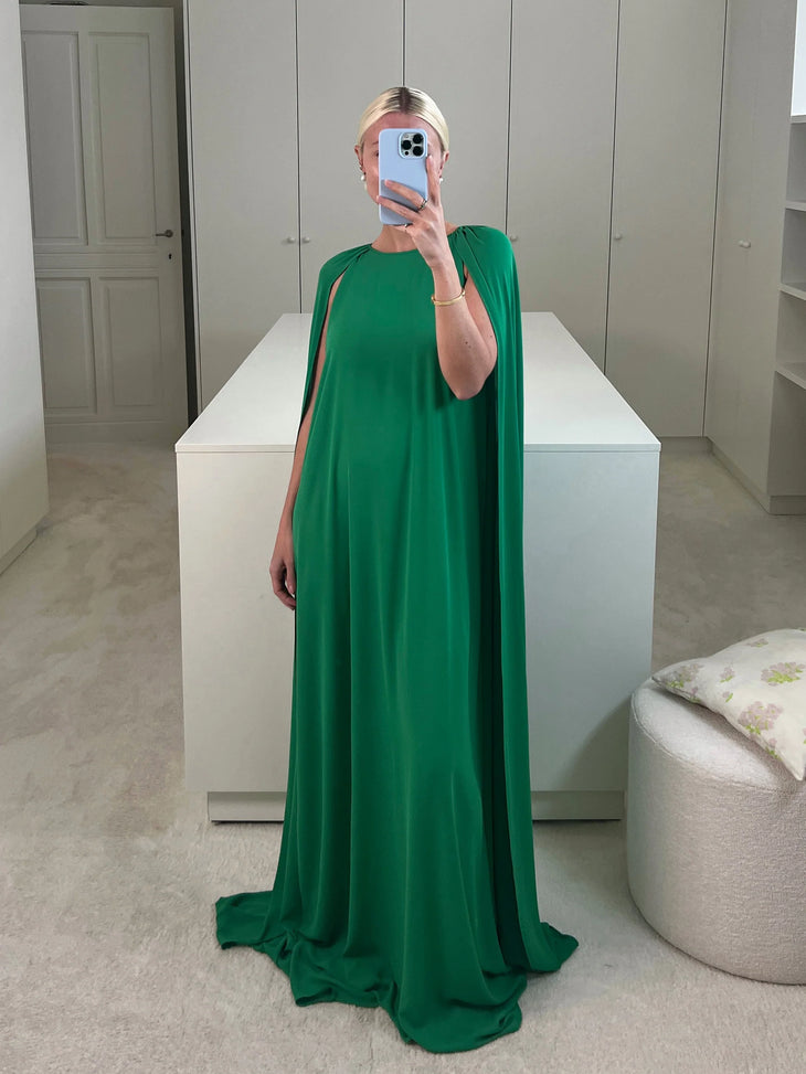 Woman wearing floor-length emerald green cape dress with flowing fabric, taking mirror selfie in modern minimalist room — Bernadette | Endless UAE