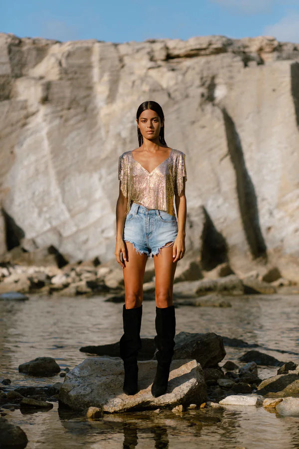 Woman wearing gold sequin Ava Top with denim shorts and black boots on rocky beach — Isabel Moore | Endless UAE