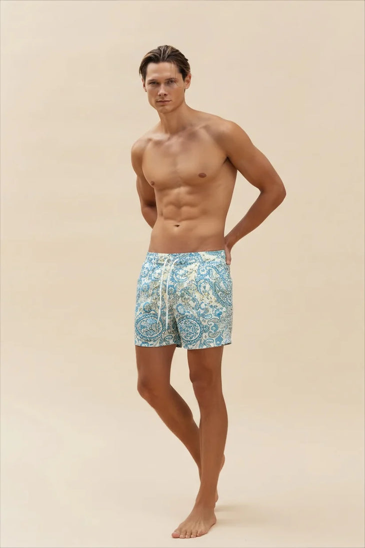Shirtless male model wearing blue and white paisley print Greek Summer swim trunks standing against beige background — Miharo | Endless UAE