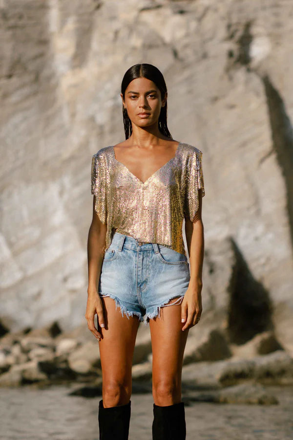 Woman wearing gold sequin Ava Top with denim shorts and black boots on rocky beach — Isabel Moore | Endless UAE