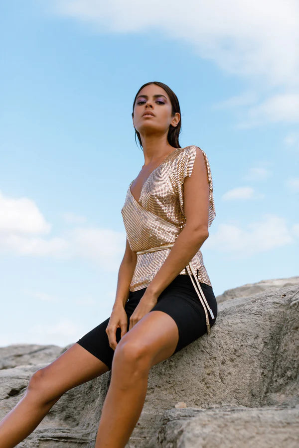 Gold sequin wrap top styled with black shorts against a rocky coastal backdrop — Isabel Moore | Endless UAE