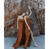 Two models wearing flowing rust-brown strapless gowns pose against textured stone backdrop — You&I Bridal | Endless UAE