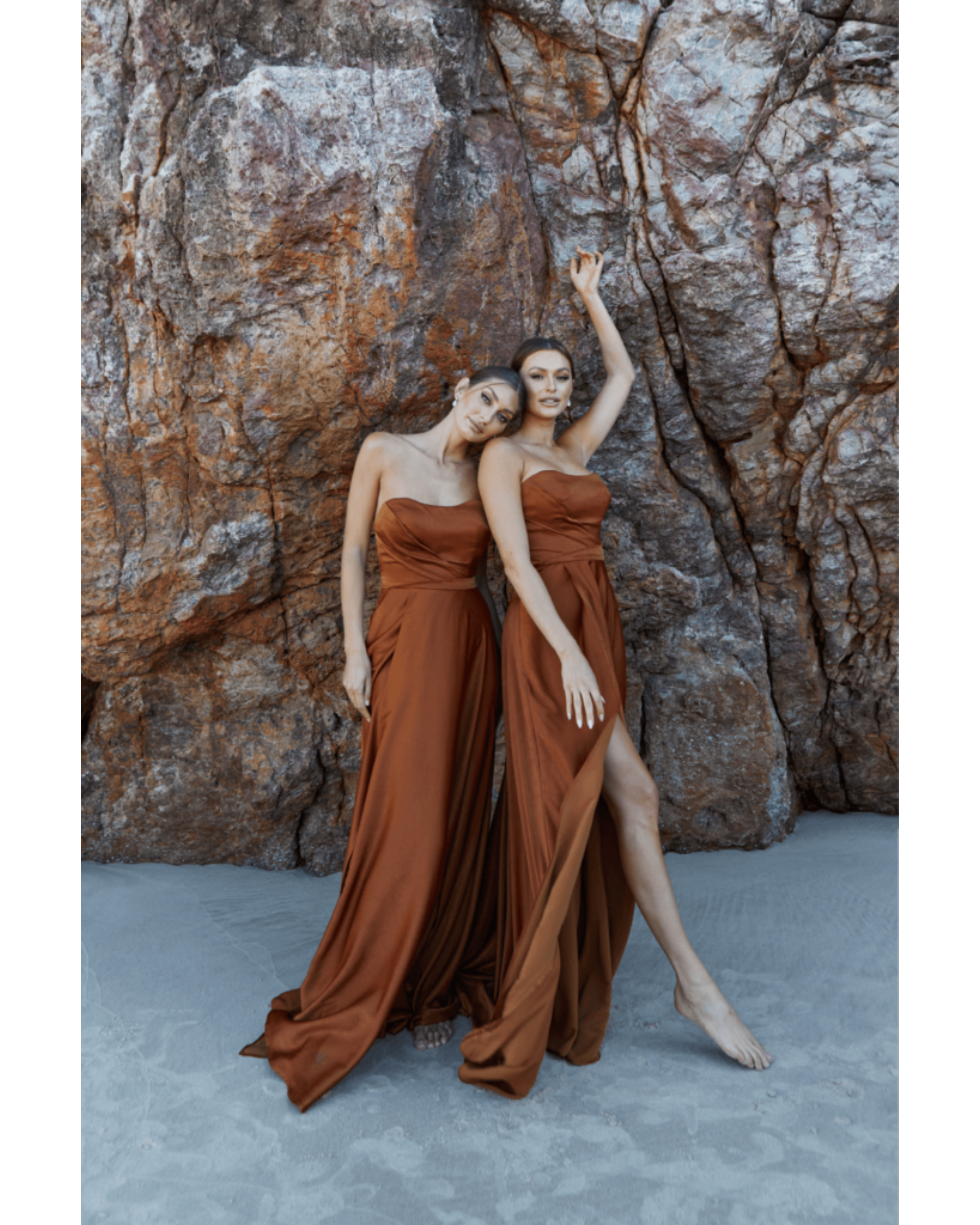 Two models wearing flowing rust-brown strapless gowns pose against textured stone backdrop — You&I Bridal | Endless UAE