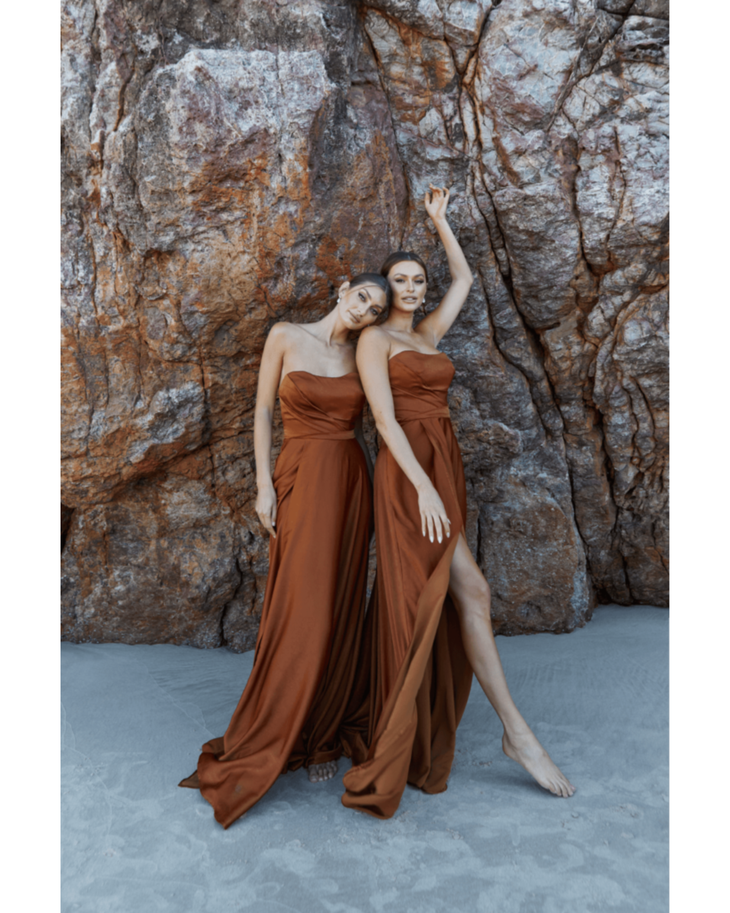Two models wearing flowing rust-brown strapless gowns pose against textured stone backdrop — You&I Bridal | Endless UAE
