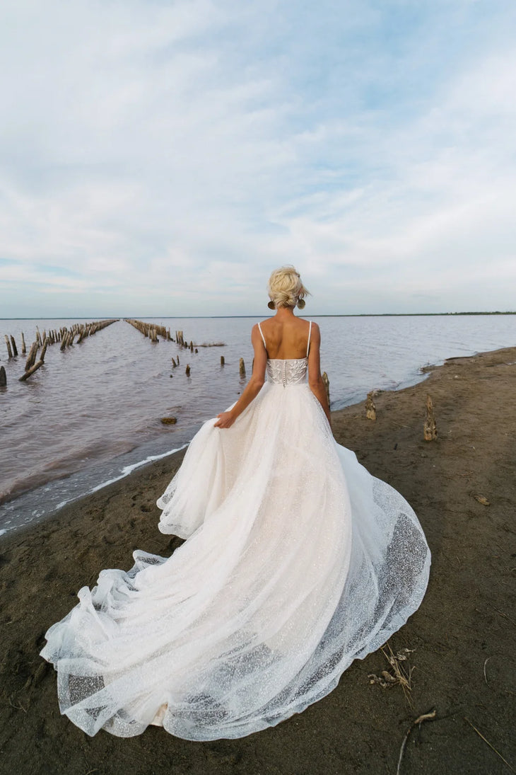 Bride in flowing white ball gown with long train standing on shore overlooking water — Romanova Atelier | Endless UAE