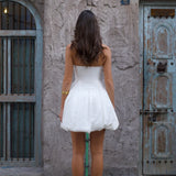 Woman in white strapless midi dress with pleated skirt and white heels against rustic wall — Cleopatress | Endless UAE