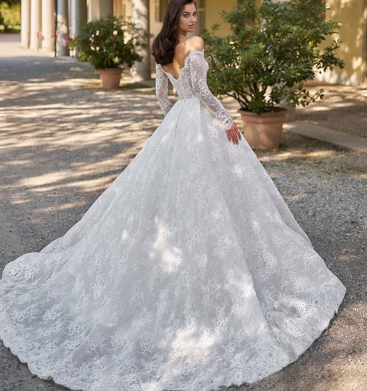 Elegant off-shoulder lace wedding gown with long sleeves and dramatic cathedral train in Italian courtyard — Sima Couture | Endless UAE