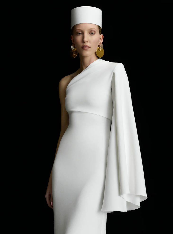 White one-shoulder maxi dress with dramatic cape sleeve and cylindrical hat, styled with gold statement earrings — Solace London | Endless UAE