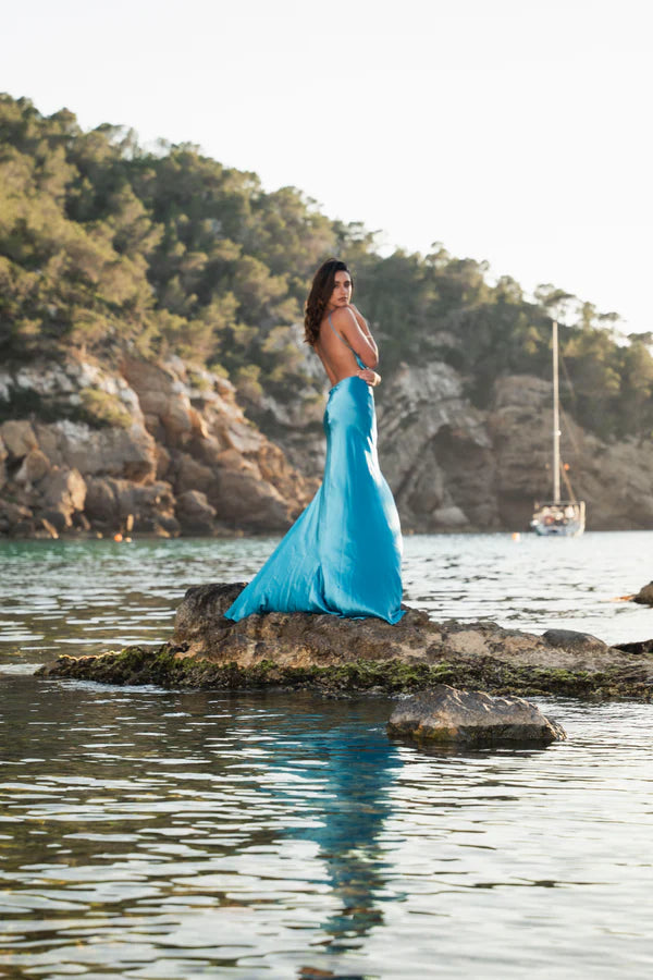Woman in turquoise backless maxi dress standing on rocks by water at sunset — By Keara | Endless UAE