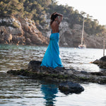 Woman in flowing turquoise backless dress standing on coastal rocks at sunset with sailboat in background — By Keara | Endless UAE