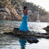 Woman in flowing turquoise backless dress standing on coastal rocks at sunset with sailboat in background — By Keara | Endless UAE