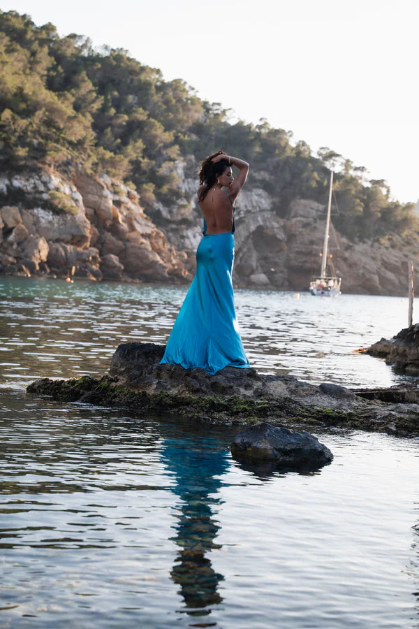 Woman in flowing turquoise backless dress standing on coastal rocks at sunset with sailboat in background — By Keara | Endless UAE