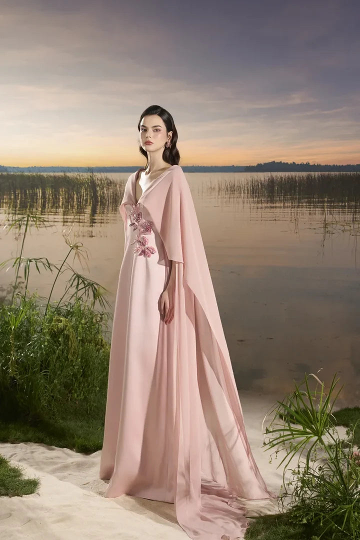 Woman wearing blush pink flowing gown with floral embellishment and draped cape at lakeside sunset — Tracy Studio | Endless UAE