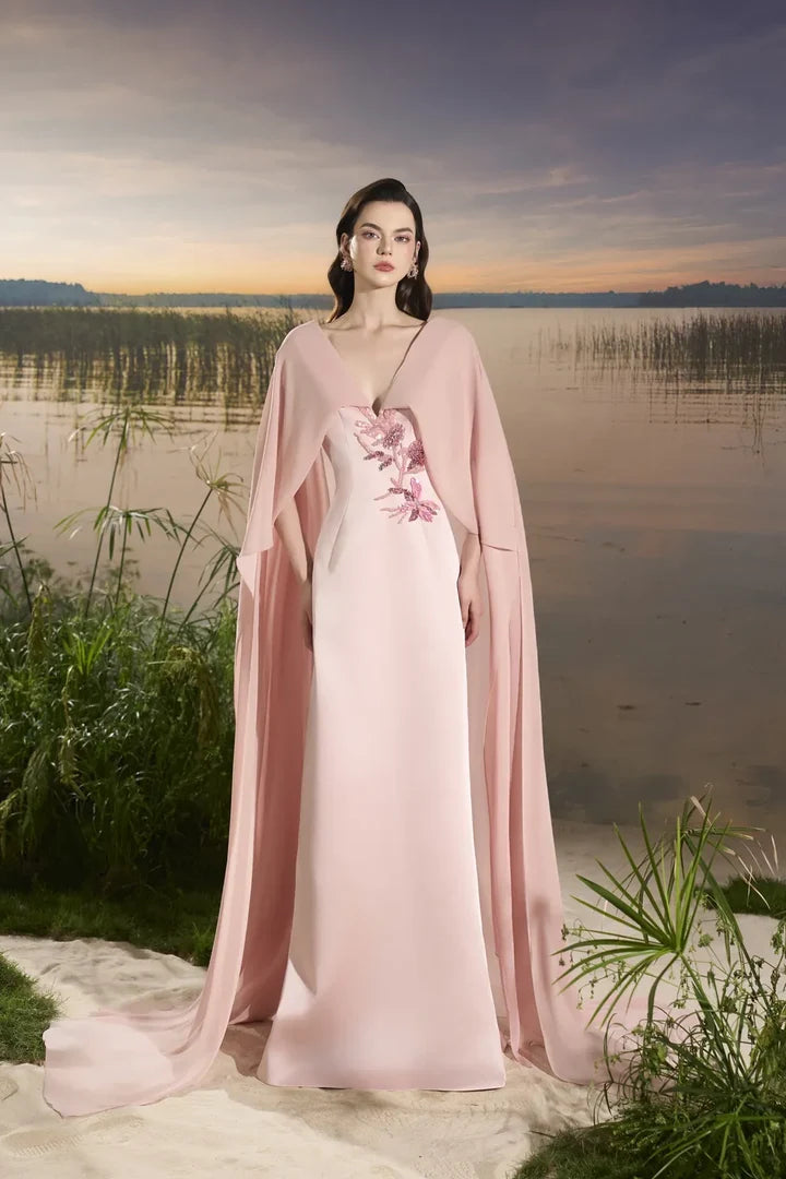 Woman wearing blush pink flowing gown with floral embellishment and draped cape at lakeside sunset — Tracy Studio | Endless UAE