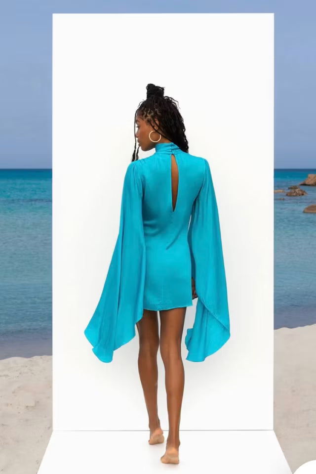 Woman wearing turquoise Farrah Dress with flowing cape sleeves at beach — Rat & Boa | Endless UAE