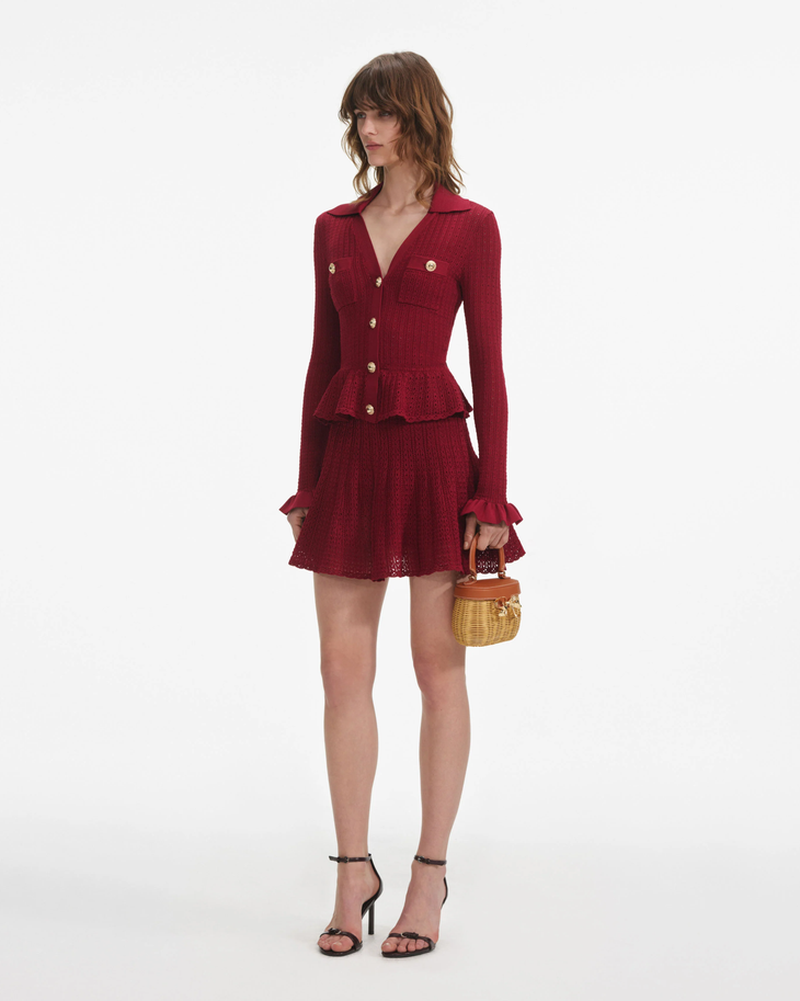 Woman wearing burgundy pointelle knit mini dress with peplum waist and ruffled cuffs, paired with black heeled sandals and woven handbag — Self-Portrait | Endless UAE