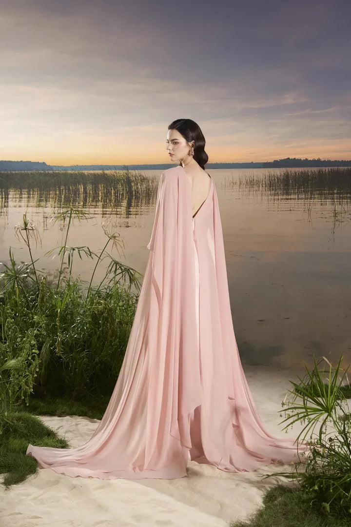Woman wearing blush pink flowing gown with floral embellishment and draped cape at lakeside sunset — Tracy Studio | Endless UAE