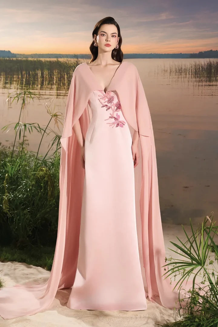 Woman wearing blush pink flowing gown with floral embellishment and draped cape at lakeside sunset — Tracy Studio | Endless UAE