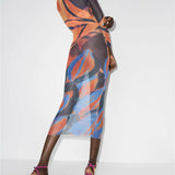 Model wearing abstract printed mesh maxi dress with long sleeves in orange, blue and purple tones, paired with pink strappy heels — Louisa Ballou | Endless UAE