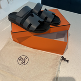 Black leather slide sandals with cutout straps displayed on orange box — Hermès | Endless UAE