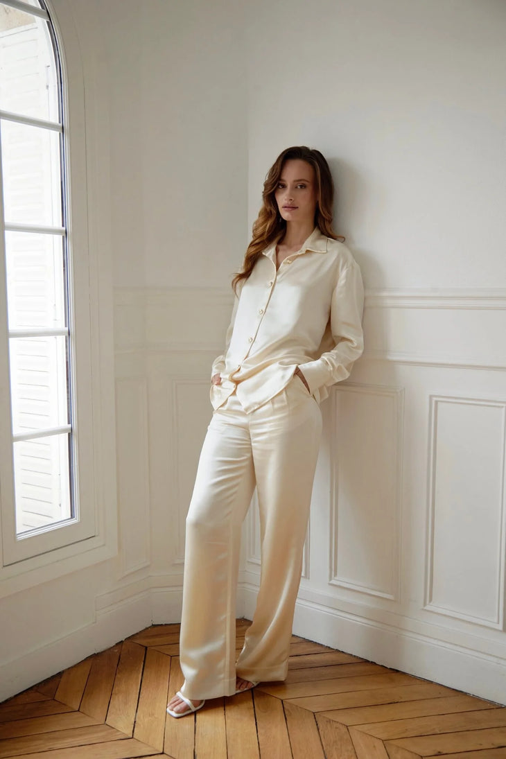 Woman wearing cream silk pajama set standing by window with herringbone floor — Nora The Label | Endless UAE