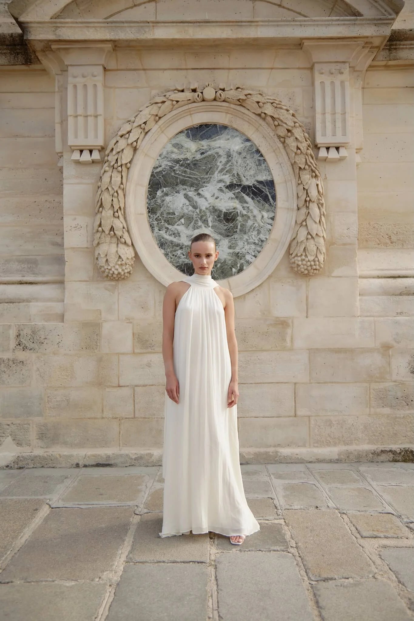 Woman wearing white flowing halter neck maxi dress standing in front of ornate stone archway — Nora The Label | Endless UAE