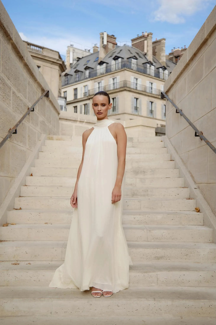 Elegant white halter neck maxi dress on model standing on Parisian staircase — Nora The Label | Endless UAE