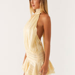 Woman wearing a pale yellow halter neck mini dress with pleated details and ruffle skirt, side view — Peppermayo | Endless UAE