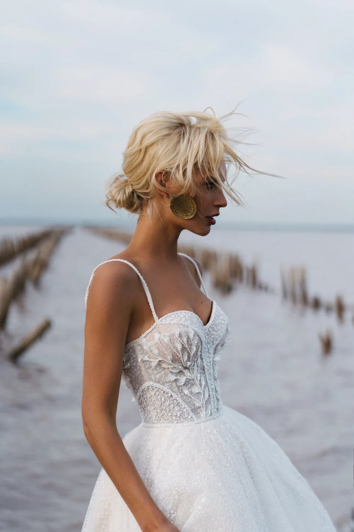 Blonde bride in beaded corset ball gown with floral appliqué by waterside, windswept hair and gold earrings — Romanova Atelier | Endless UAE