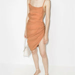 Peach asymmetrical mini dress with ruched side detail and thin straps on model with white bag and strappy heels — Jacquemus | Endless UAE