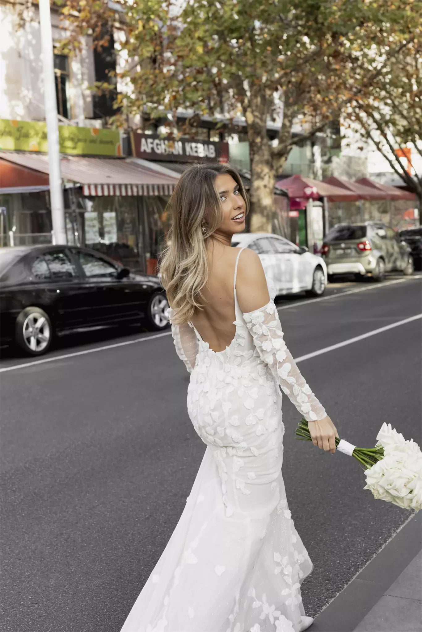 Elegant fitted wedding dress with floral appliqué sleeves and backless design, bride holding white bouquet on city street — You&I Bridal | Endless UAE