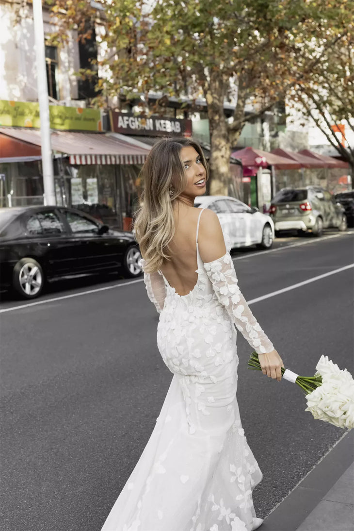 Elegant fitted wedding dress with floral appliqué sleeves and backless design, bride holding white bouquet on city street — You&I Bridal | Endless UAE
