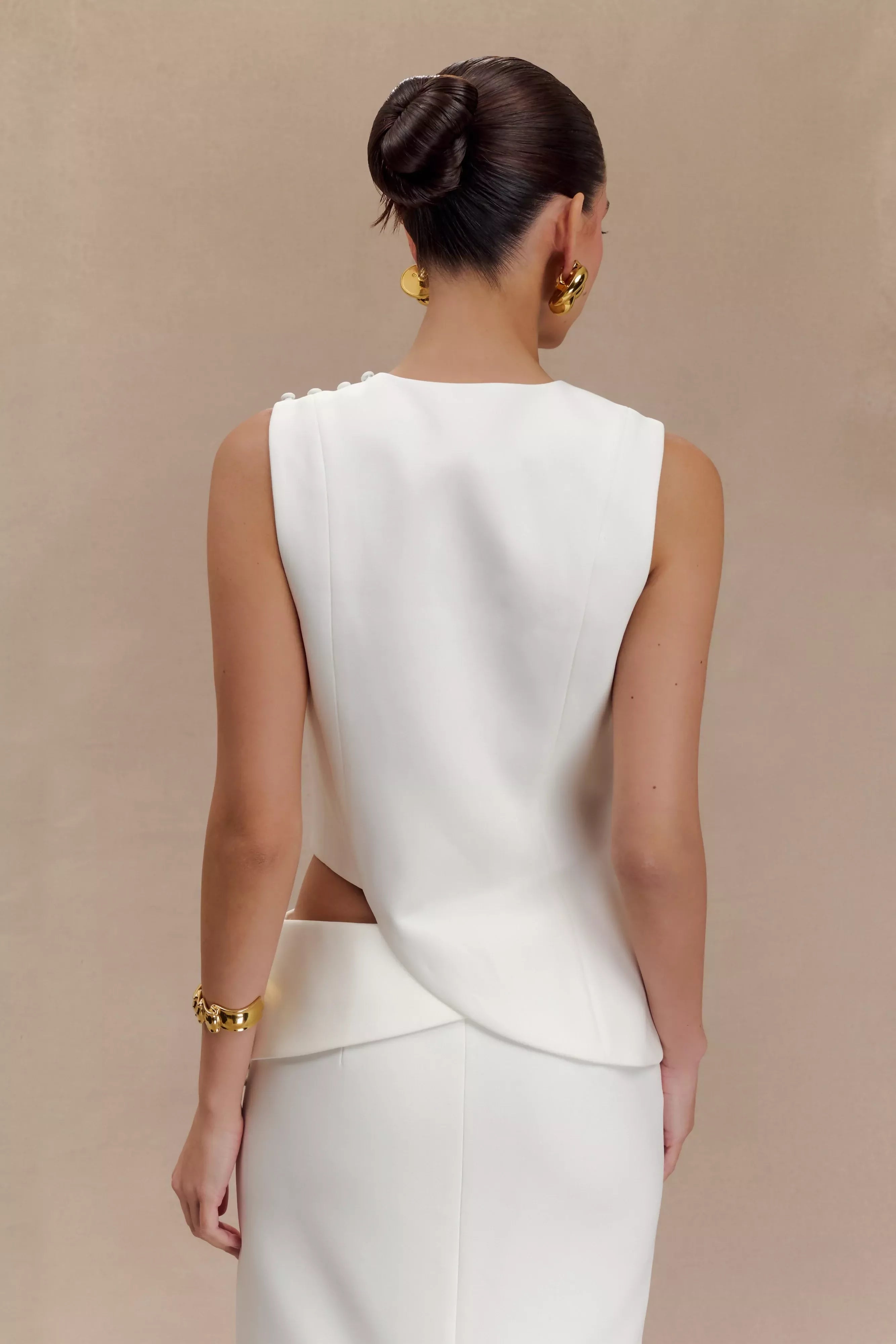 Rear view of woman wearing white sleeveless asymmetric suiting top with gold jewelry and hair in bun — Endless | Endless UAE