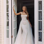 Bride in fitted lace wedding gown with illusion neckline and flowing tulle overskirt standing in white doorway — You&I Bridal | Endless UAE
